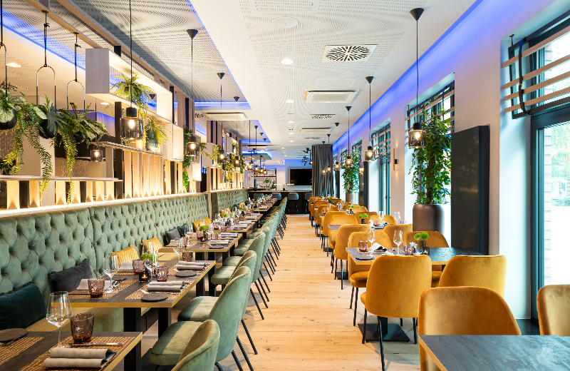 A modern dining room with green banquette seating, yellow chairs, and hanging lights/plants.