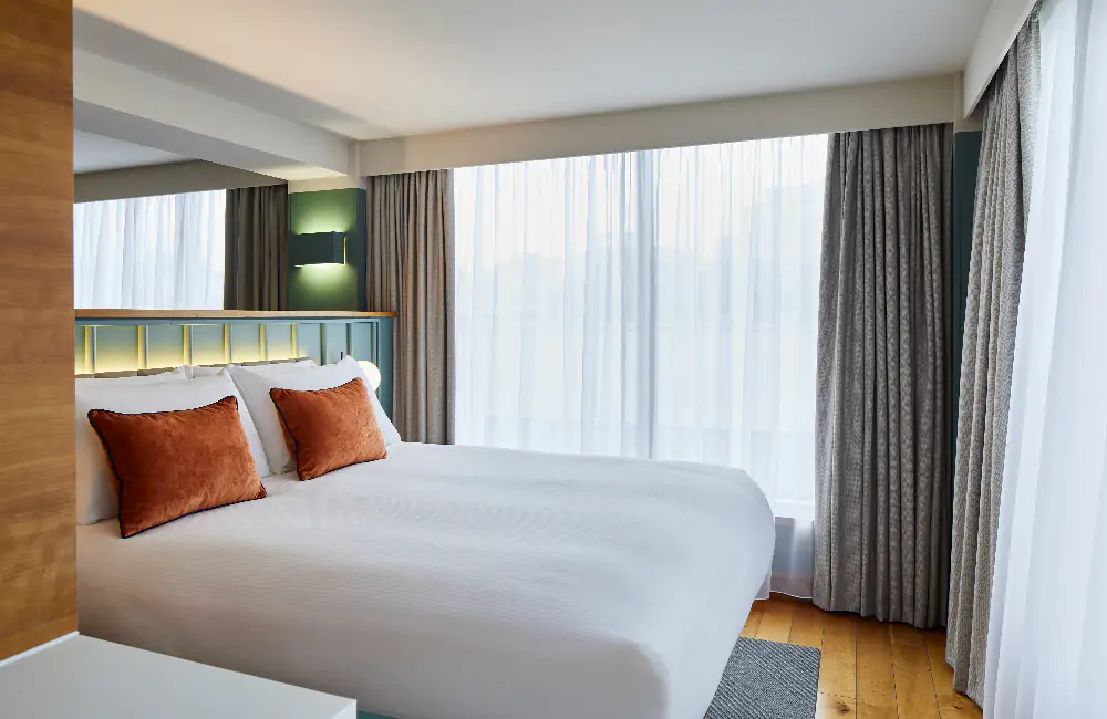 Bright bedroom at master Farringdon London serviced apartments, featuring a large double bed with crisp white linens and two orange accent pillows, framed by floor-to-ceiling windows and soft natural light.