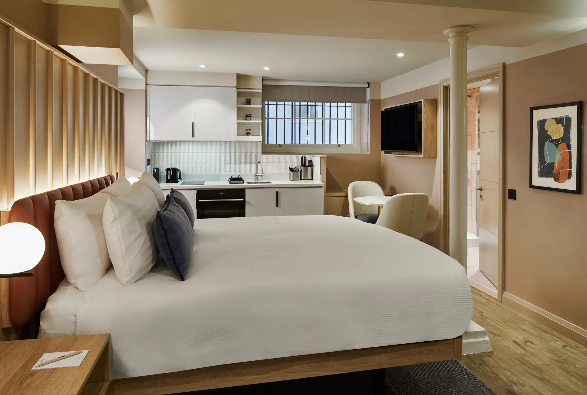 Cozy lower ground studio at master Cannon London featuring a large bed, fully-equipped kitchenette, small dining area, and natural light from a high window.