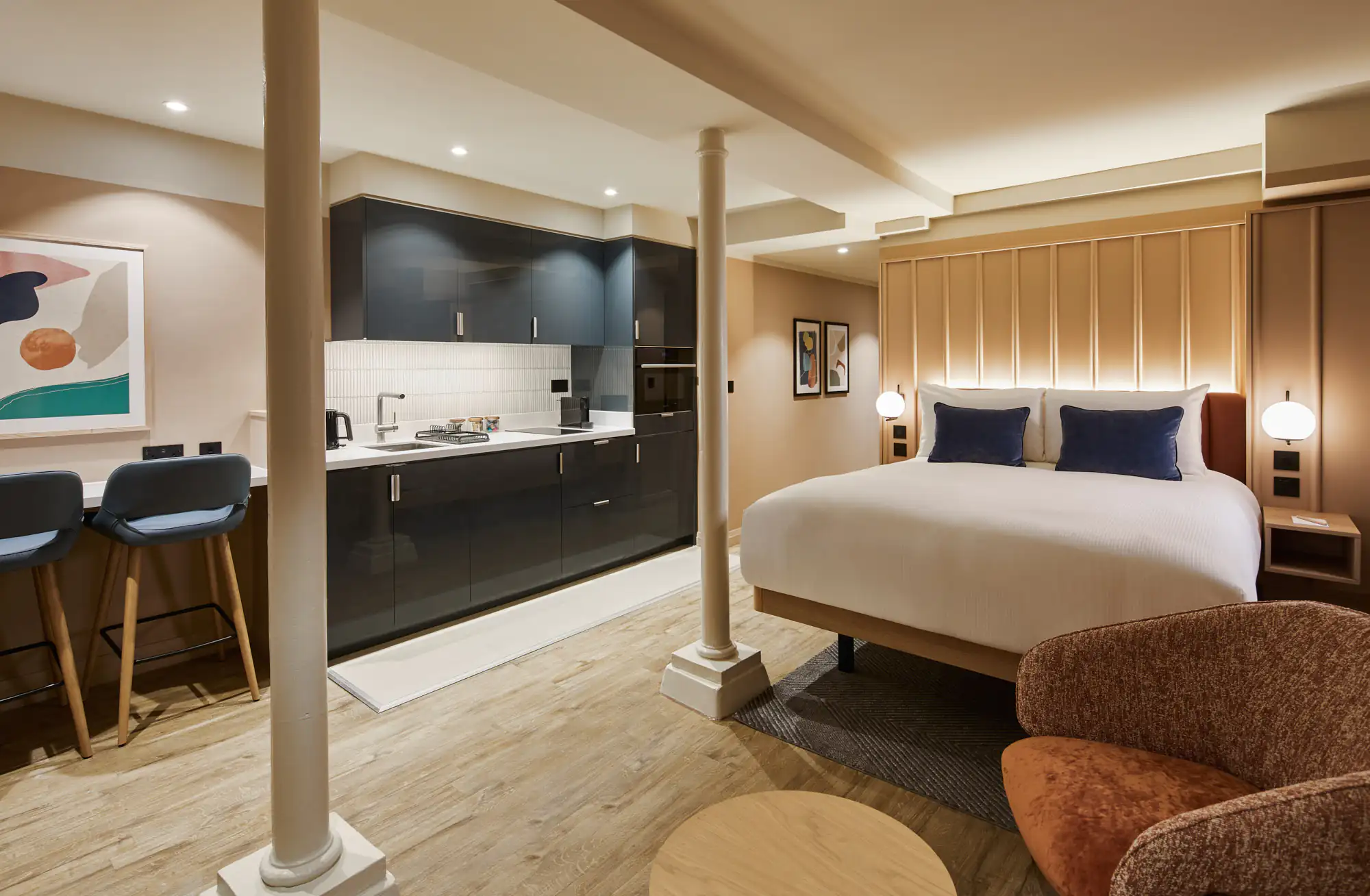 Spacious lower ground studio at master Cannon London combining a stylish bed, sleek dark kitchen units with bar seating, and warm ambient lighting.