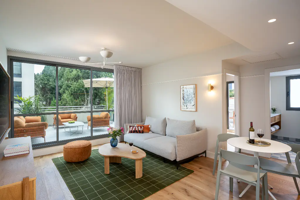 Bright and stylish living room in master Mazeh apartment featuring a light grey sofa, green checkered rug, and large glass doors opening to a spacious terrace with outdoor seating.
